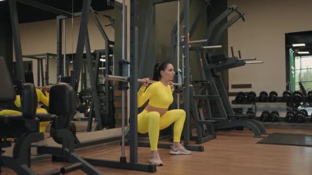 Spor salonunda ya da fitness kulübünde Inflight Fitness Smith Machine kullanarak vücut kasları geliştiren İspanyol genç kadın. Kız omuzlarında halterle ağırlık kaldırırken, çömelmeler sağlık için egzersiz yapıyor.. — Stok video