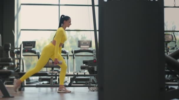 Молодая мускулистая латиноамериканка делает выпады с гантелями в спортзале в жёлтой спортивной одежде. Женщина-спортсмен с гантелями в положении выпада в тренажерном зале. — стоковое видео