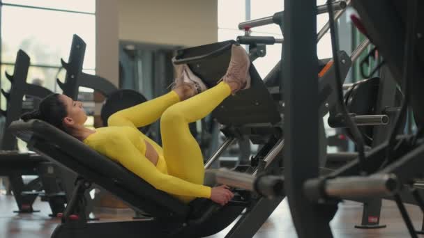 Mujer hispana usando una máquina de prensa en un club de fitness. Mujer joven haciendo un ejercicio sobre sus pies en el simulador en ropa deportiva amarilla, — Vídeo de stock