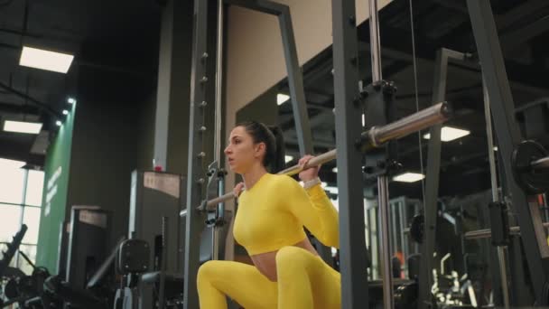 トレーニングジムやフィットネスクラブでフライトフィットネススミスマシンを使用して体の筋肉を構築するヒスパニック系の若い女性。女の子は肩の上にベルで体重を持ち上げる一方、健康のためにスクワット運動. — ストック動画
