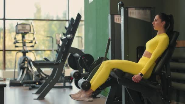 Sterke latino brunette sportvrouw doet beentraining op sport simulator in fitness club. In gele sportkleding. atletische vrouw voert een uitbreiding been oefening in de simulator — Stockvideo