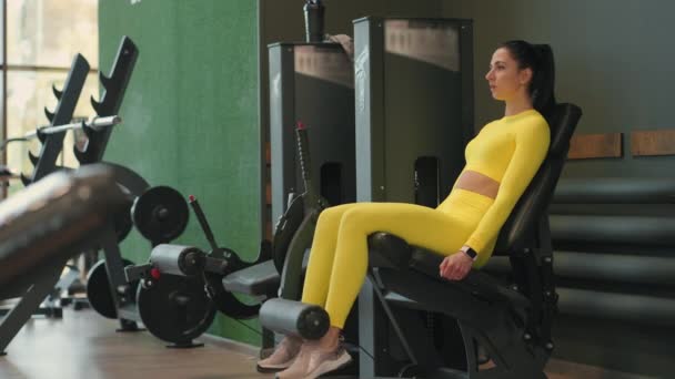Forte sportive brune hispanique faisant des exercices de jambes sur simulateur de sport dans un club de fitness. En tenue de sport jaune. Femme sportive effectue un exercice de jambe d'extension dans le simulateur — Video