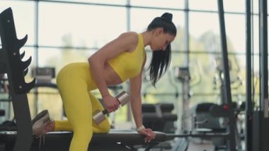 Spor giyim antrenmanındaki İspanyol atlet kadın bir elinde dambılla spor salonundaki tezgaha yaslanıyor.