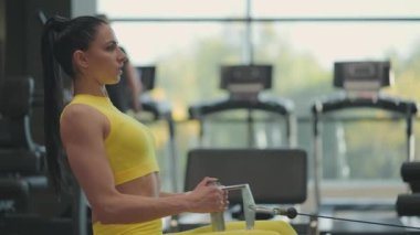 İspanyol kadın kürek çekme makinesinde egzersiz yapıyor. Simülatörün arkasında oturan bir kıza kürek çekme makinesi denir. Oturma ağırlığı büyük pencereler ve spor salonunun koşu bantları kadar.