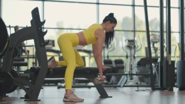 Spor giyim antrenmanındaki İspanyol atlet kadın bir elinde dambılla spor salonundaki tezgaha yaslanıyor.