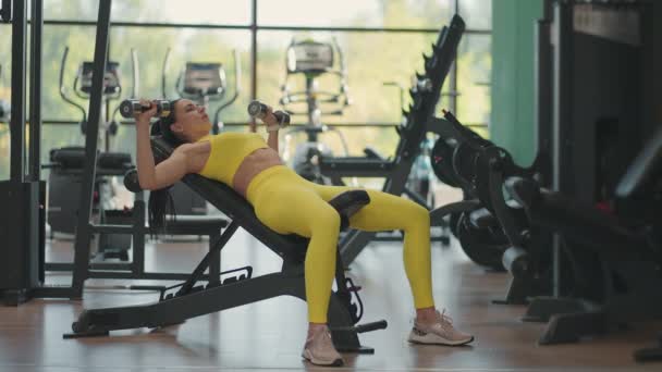 Beslutsam latinamerikansk kvinnlig idrottsman gör bänk press med hantlar i gymmet. En vältränad ung kvinna som gör en hantelbänkspress i ett litet gym. Muskulös kvinnlig kroppsbyggare liggande bänk och gör hantel — Stockvideo