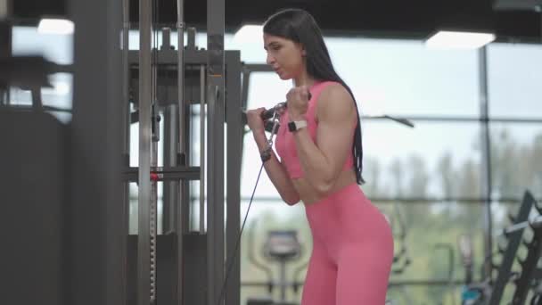 분홍색 양복을 입은 갈색 머리의 여자가 이두근 훈련을 위해 교차하는 팔로 밧줄을 잡아당긴다. 훈련을 받도록 하 십시오. 전문 여성 강사 — 비디오