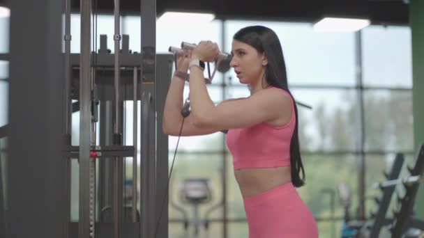 En latinamerikansk brunette i lyserødt jakkesæt trækker et reb i en crossover med hænderne for at træne sine skuldre. Skulder træning i en træner. Professionel kvinde instruktør – Stock-video
