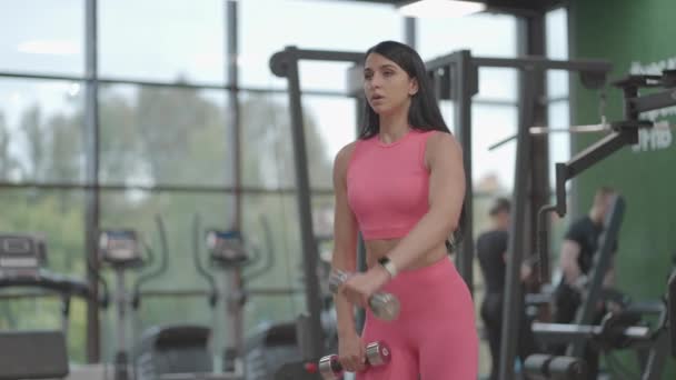 Uma mulher morena em um terno rosa alternadamente levanta halteres para o topo na frente dela enquanto treina os ombros no ginásio. Exercício de pé para treinar os ombros e braços. Treinamento de peso — Vídeo de Stock