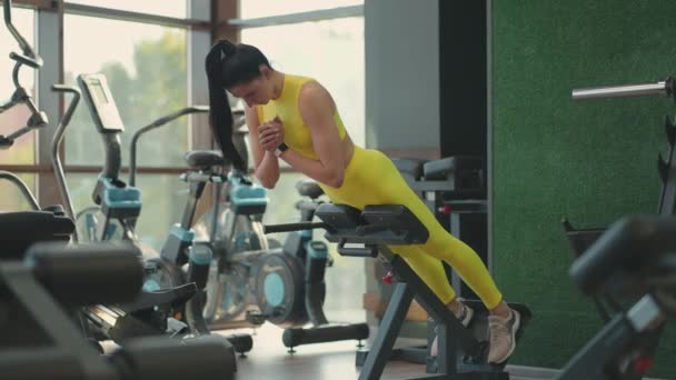 Jovem mulher hispânica em um traje amarelo realiza um exercício de hiperextensão para treinar os músculos das costas. Uma mulher morena levanta o corpo no banco do ginásio para fortalecer as costas e a postura — Vídeo de Stock