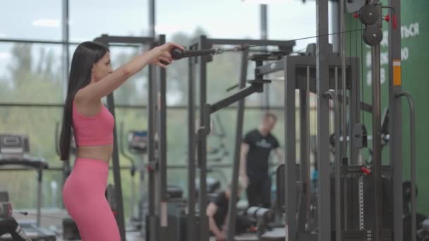 Una donna bruna in un abito rosa tira una corda in un allenatore crossover al petto. Allenamento della schiena in un allenatore. Professionale donna istruttrice — Video Stock