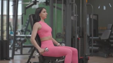 Pembe takım elbiseli esmer bir kadın spor salonunda omuzlarını çalıştırırken halterlerini yana kaldırıyor. Oturmuş Omuz ve Kol Yedeği Egzersizi
