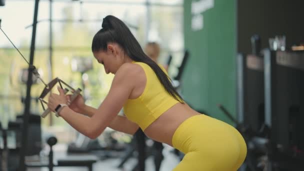 Una joven hispana en chándal amarillo realiza un ejercicio en un crossover tira de una cuerda de acero desde arriba para entrenar su espalda y hombros. Una mujer entrena su espalda y hombros en un gimnasio — Vídeos de Stock