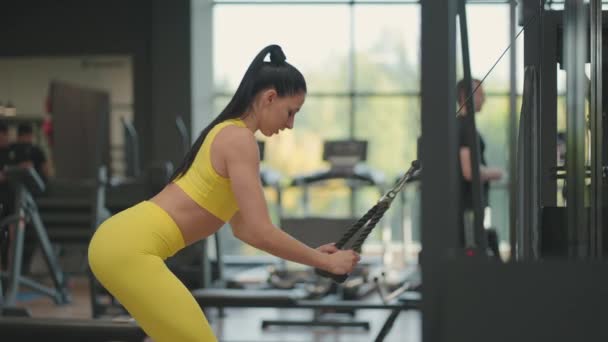 Una joven hispana en chándal amarillo realiza un ejercicio en un crossover tira de una cuerda desde arriba para entrenar la espalda y los hombros. Mujer morena entrena espalda y hombros en el gimnasio — Vídeos de Stock