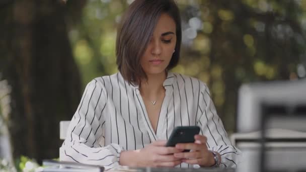 Jonge Latijns-Amerikaanse etnische vrouw zit in een zomer cafe met telefoon kijken naar het scherm kijken naar sociale media video-inhoud, film of streamen online in mobiele app lachen met behulp van een smartphone — Stockvideo