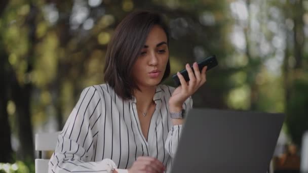 Multitasking hispanic businesswoman using voice recognition function on a smartphone standing near desk and typing on the laptop. Arabian female entrepreneur sending a vocal message — Stock Video
