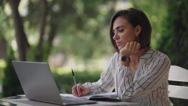 Estudante Mulher Morena árabe ethnos hispânicos estudando remotamente através da Internet, enquanto sentado em um café de verão com um laptop e escrever uma caneta e um caderno ao tomar notas no webinar — Vídeo de Stock