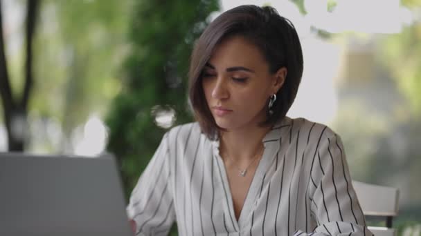 Studente Donna Bruna Arabo Ispanico ethnos studiare a distanza via Internet mentre seduto in un caffè estivo con un computer portatile e scrivere una penna e un taccuino mentre prende appunti sul webinar — Video Stock