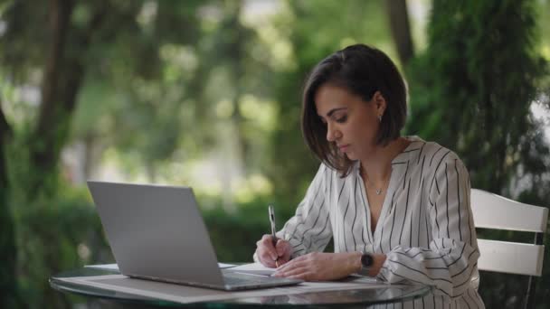 Estudiante mujer morena árabe hispano ethnos estudiar de forma remota a través de Internet mientras está sentado en un café de verano con un ordenador portátil y escribir un bolígrafo y un cuaderno mientras toma notas en el webinar — Vídeos de Stock