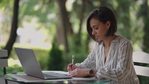 Donna d'affari Bruna Arabo Ispanico Etnia Lavora a distanza mentre è seduto in un caffè estivo in una giornata di sole con un computer portatile e scrive con una penna e un quaderno — Video Stock