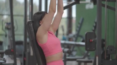 Pembe takım elbiseli esmer bir kadın spor salonunda omuzlarını eğitirken dambılı yukarı kaldırıyor. Kollarını ve omuzlarını çalıştırmak için bir bankta oturarak egzersiz yap. Ağırlık çalışması