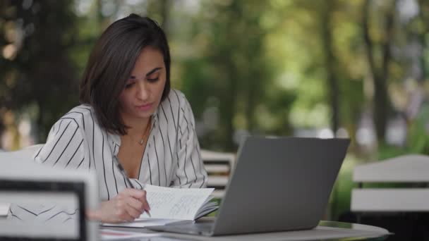 Mulher de negócios Morena Árabe Hispânica Etnia Funciona remotamente enquanto está sentado em um café de verão em um dia ensolarado com um laptop e escreve com uma caneta e caderno — Vídeo de Stock