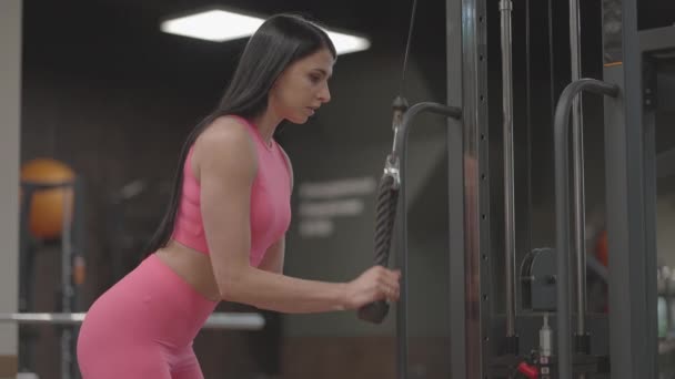 A brunette woman in a pink suit pulls a rope in a crossover arm for triceps training. Hand training in a trainer. Professional woman instructor — Stock Video