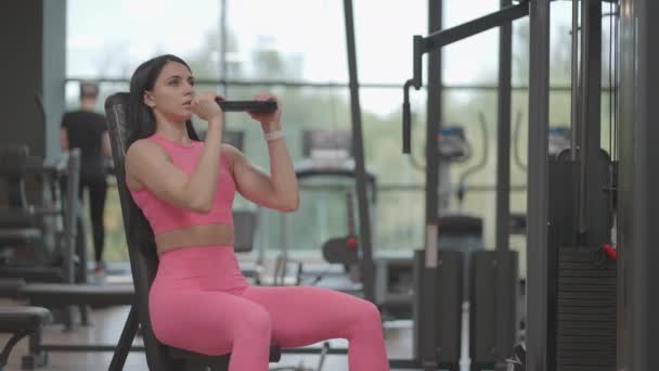 Pembe takım elbiseli esmer bir kadın spor salonunda omuzlarını eğitirken dambılı yukarı kaldırıyor. Kollarını ve omuzlarını çalıştırmak için bir bankta oturarak egzersiz yap. Ağırlık çalışması — Stok video
