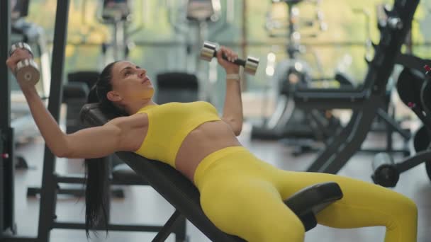 Belle femme sportive entraîne les muscles pectoraux en élevant des haltères tout en étant couché dans la salle de gym. Entraînement musculaire féminin d'athlète avec haltères, soulevant des poids — Video