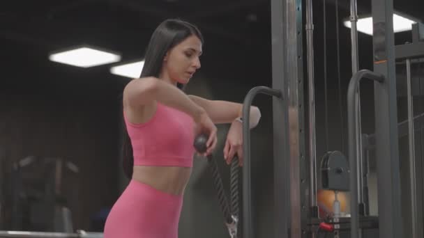 A hispanic brunette woman in a pink suit pulls a rope in a crossover with her hands to train her shoulders. Shoulder workout in a trainer. Professional woman instructor — Stock Video