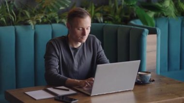 Gömlekli genç bir adam elinde dizüstü bilgisayarla bir masada oturuyor ve klavyeye yazı yazıyor. Bir öğrenci uzaktan çalışabilir. Bir iş adamı işini uzaktan yürütür.