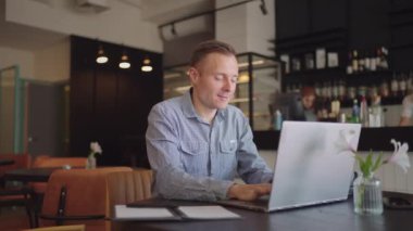 Gömlekli genç bir adam elinde dizüstü bilgisayarla bir masada oturuyor ve klavyeye yazı yazıyor. Bir öğrenci uzaktan çalışabilir. Bir iş adamı işini uzaktan yürütür.