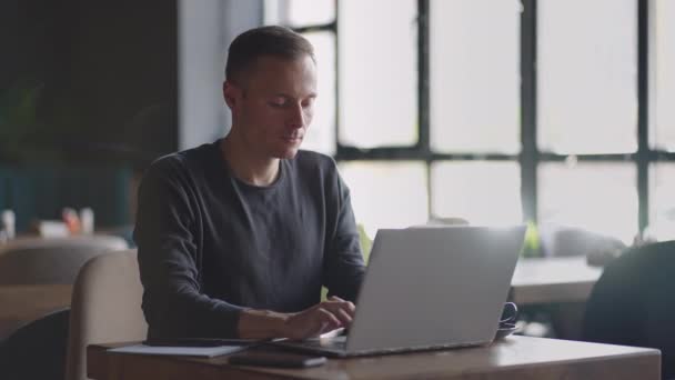 Um jovem de camisa está sentado em uma mesa com um laptop e digitando no teclado. Um estudante pode estudar remotamente. Um homem de negócios conduz o seu negócio remotamente — Vídeo de Stock