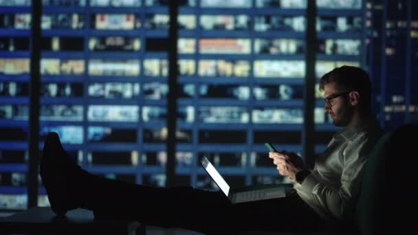 Um homem deitado em um sofá com um laptop no colo e escrevendo uma mensagem em um telefone celular com vista para a cidade noturna em casa — Vídeo de Stock