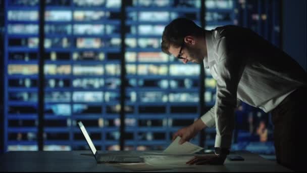 Un hombre barbudo con gafas en el fondo de la ventana de una gran ciudad nocturna trata con informes y análisis de proyectos de valores y pronósticos de negocios. — Vídeos de Stock