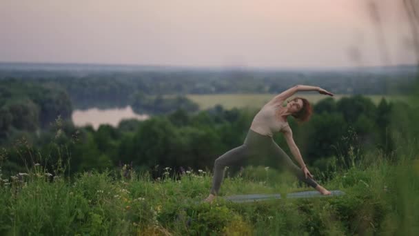 En ung kvinna håller balansen genom att göra yogaövningar på en matta i naturen i bakgrunden en fallskärmshoppare flyger i slow motion — Stockvideo