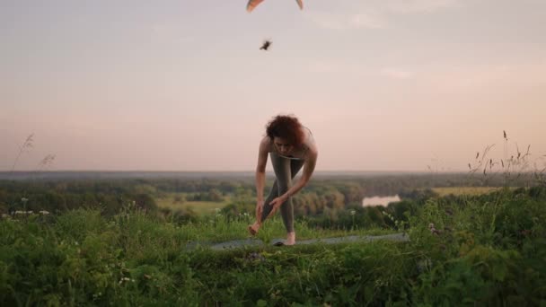Μια νεαρή γυναίκα κρατά την ισορροπία της κάνοντας ασκήσεις γιόγκα σε ένα χαλί στη φύση στο παρασκήνιο ένας αλεξιπτωτιστής αλεξίπτωτο πλαγιάς πετάει σε αργή κίνηση — Αρχείο Βίντεο