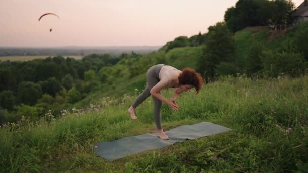 Mladá žena udržuje rovnováhu tím, že cvičí jógu na koberci v přírodě v pozadí parašutista létá ve zpomaleném filmu — Stock video