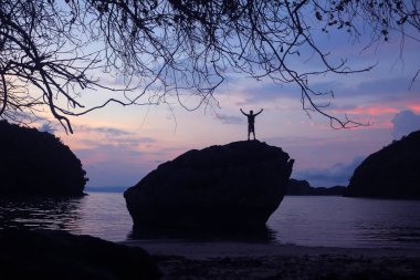 Sahildeki bir kayanın tepesinde gün doğumunda, sabah karanlığı çökmüş bir adam..
