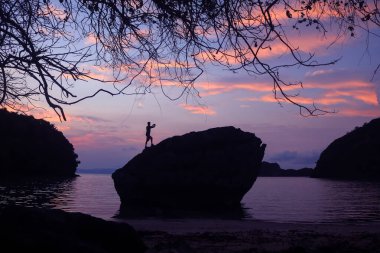 Bir adam sabah güneşinin fotoğrafını çekmek için telefon kullanıyor. Sahildeki bir kayanın tepesinde, güneş doğarken, gökyüzü karartması..