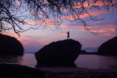 Bir adam sabah güneşinin fotoğrafını çekmek için telefon kullanıyor. Sahildeki bir kayanın tepesinde, güneş doğarken, gökyüzü karartması..