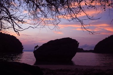 Sahildeki bir kayanın tepesinde gün doğumunda, sabah karanlığı çökmüş bir adam..