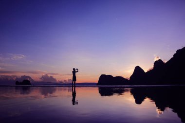 Sahilde güneşin doğuşunu fotoğraflamak için telefon kullanan adam su, deniz ve dağ arkasını yansıtıyor..