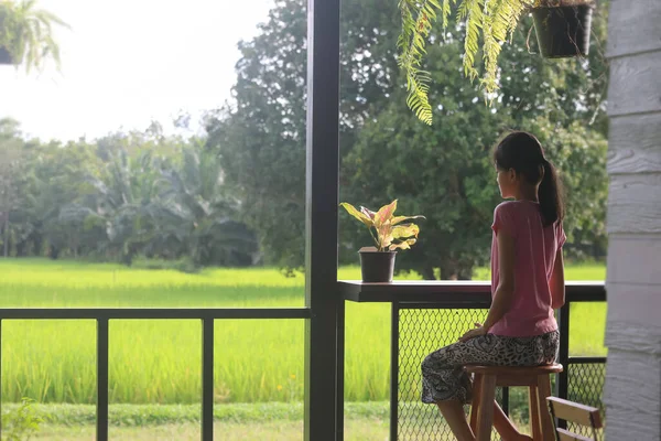 An Asian girl sits on a chair on the balcony, looking at the fields and nature