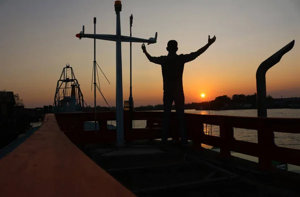 An Asian man watches the sunset on a fishing boat parked by a river in Thailand.