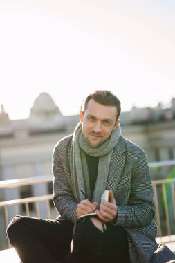 Eşarplı ve ceketli genç, gülümseyen sarışın adam kameraya bakıyor, notla oturuyor ve yazıyor. Açık hava Avrupa kenti çalışma konsepti.