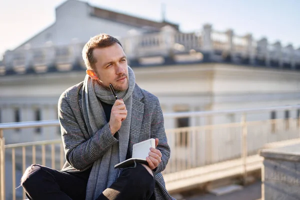 Attractive man with beard in gray scarf and coat sitting on the roof and thinking, holding notes and pen. European city concept of working outdoor.