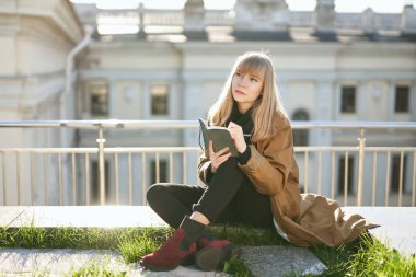 Genç çekici sarışın kız oturuyor, düşünüyor ve not defterini yeşil çimlere yazıyor. Açık hava çalışma konsepti