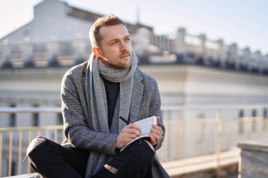 Gri atkı ve ceketli genç sakallı erkek çatıda oturmuş, elinde not defteri ve kalemle düşünüyor. Açık hava çalışma konsepti