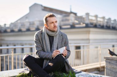 Çatıda oturan gri atkılı ve ceketli genç adam elinde notlar ve kalemle düşünüyor.